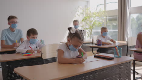 Multiracial-group-of-kids-wearing-face-masks-working-at-class-writing-and-listening-explanations-of-teacher-in-classroom